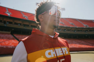 1UP Sports Marketing client Patrick Mahomes standing in a Chiefs sweatshirt and sunglasses with sun's rays coming through